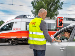 Ein Mann in ÖBB-Sicherheitsweste vor einem Bahnübergang reicht einer Frau im Auto einen Informationsflyer. Das Auto steht an einer roten Ampel an einem Bahnübergang. Ein ÖBB-Zug fährt gerade durch.