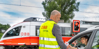 Ein Mann in ÖBB-Sicherheitsweste vor einem Bahnübergang reicht einer Frau im Auto einen Informationsflyer. Das Auto steht an einer roten Ampel an einem Bahnübergang. Ein ÖBB-Zug fährt gerade durch.
