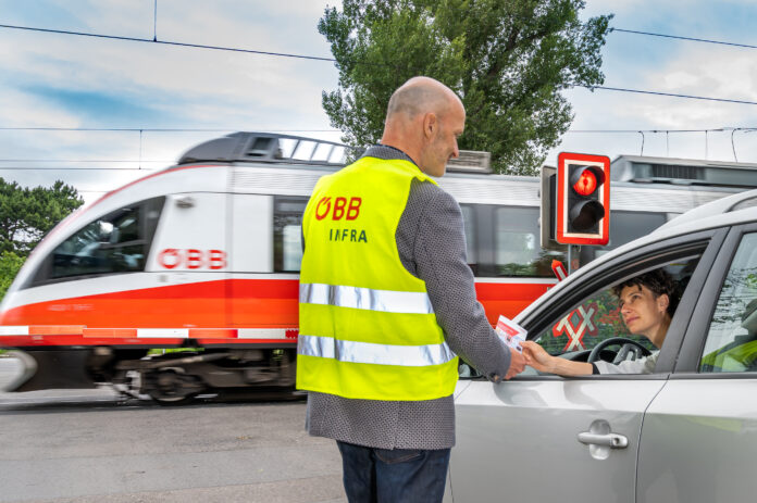 Eisenbahnkreuzung_c_OEBB_Michael Fritscher Ein Mann in ÖBB-Sicherheitsweste vor einem Bahnübergang reicht einer Frau im Auto einen Informationsflyer. Das Auto steht an einer roten Ampel an einem Bahnübergang. Ein ÖBB-Zug fährt gerade durch.
