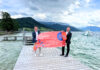 Zwei Männer halten orange Schwimmbojen und ein Plakat hoch, das den Ablauf des Ausleihens von Schwimmbojen erklärt. Sie stehen auf einem Steg am Attersee.