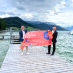 Zwei Männer halten orange Schwimmbojen und ein Plakat hoch, das den Ablauf des Ausleihens von Schwimmbojen erklärt. Sie stehen auf einem Steg am Attersee.
