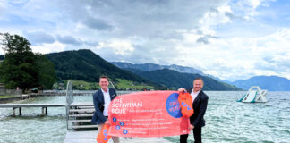 Zwei Männer halten orange Schwimmbojen und ein Plakat hoch, das den Ablauf des Ausleihens von Schwimmbojen erklärt. Sie stehen auf einem Steg am Attersee.