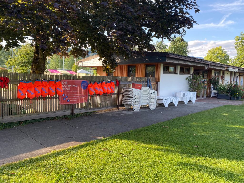 An einem Zaun eines Badehäuschens sind aufgeblasene orange-farbene Schwimmbojen mit KFV-Logo aufgehängt. Auch eine Anleitung des Gratis-Verleihs auf einem Banner ist angebracht.
