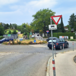 Drei dunkle Autos in einem Kreisverkehr. Davor ein Vorrang-Schild und der Hinweis "Blinken beim Verlassen".