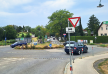 Drei dunkle Autos in einem Kreisverkehr. Davor ein Vorrang-Schild und der Hinweis "Blinken beim Verlassen".