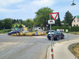 Drei dunkle Autos in einem Kreisverkehr. Davor ein Vorrang-Schild und der Hinweis "Blinken beim Verlassen".