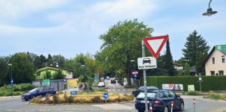 Drei dunkle Autos in einem Kreisverkehr. Davor ein Vorrang-Schild und der Hinweis "Blinken beim Verlassen".