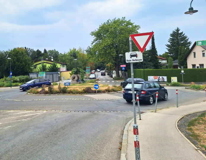 Drei dunkle Autos in einem Kreisverkehr. Davor ein Vorrang-Schild und der Hinweis "Blinken beim Verlassen".