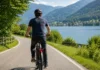 Radfahrer auf einer Landstraße entlang eines Sees mit Bergen im Hintergrund