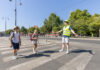 Zwei Kinder im Volksschulalter gehen über einen Zebrastreifen. Dabei hilft ihnen ein junger Mann mit Warnweste und Signalstab sicher über die Straße. Im Hintergrund sieht man die Ringstraße und Bäume. Die Sonne schein und es ist keine Wolke am Himmel zu sehen.