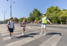Zwei Kinder im Volksschulalter gehen über einen Zebrastreifen. Dabei hilft ihnen ein junger Mann mit Warnweste und Signalstab sicher über die Straße. Im Hintergrund sieht man die Ringstraße und Bäume. Die Sonne schein und es ist keine Wolke am Himmel zu sehen.