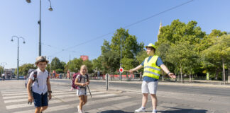 Zwei Kinder im Volksschulalter gehen über einen Zebrastreifen. Dabei hilft ihnen ein junger Mann mit Warnweste und Signalstab sicher über die Straße. Im Hintergrund sieht man die Ringstraße und Bäume. Die Sonne schein und es ist keine Wolke am Himmel zu sehen.
