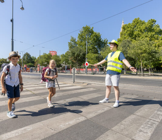 Zwei Kinder im Volksschulalter gehen über einen Zebrastreifen. Dabei hilft ihnen ein junger Mann mit Warnweste und Signalstab sicher über die Straße. Im Hintergrund sieht man die Ringstraße und Bäume. Die Sonne schein und es ist keine Wolke am Himmel zu sehen.
