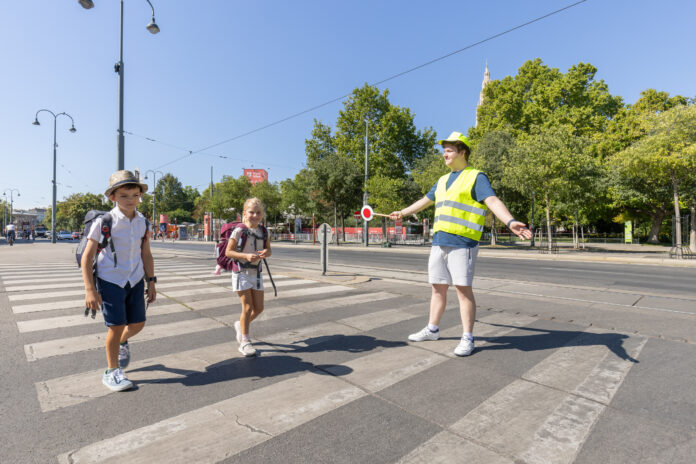 Fototermin Schülerlotsen Zwei Kinder im Volksschulalter gehen über einen Zebrastreifen. Dabei hilft ihnen ein junger Mann mit Warnweste und Signalstab sicher über die Straße. Im Hintergrund sieht man die Ringstraße und Bäume. Die Sonne schein und es ist keine Wolke am Himmel zu sehen.
