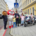 Eine Gruppe an Vertreter*innen der Polizei (Landespolizeivizepräsident General Dieter Csefan, interimistische Leiterin der Landesverkehrsabteilung Wien, Oberst Andrea Anders), Klaus Robatsch vom KFV, 3 Schulkindern mit Plüsch-HELMIS (1 davon auf einem Polizei-Motorrad) sowie den Maskottchen HELMI und dem Polizeibären vor einem Schild "Achtung Schulweg".