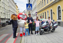 Eine Gruppe an Vertreter*innen der Polizei (Landespolizeivizepräsident General Dieter Csefan, interimistische Leiterin der Landesverkehrsabteilung Wien, Oberst Andrea Anders), Klaus Robatsch vom KFV, 3 Schulkindern mit Plüsch-HELMIS (1 davon auf einem Polizei-Motorrad) sowie den Maskottchen HELMI und dem Polizeibären vor einem Schild "Achtung Schulweg".