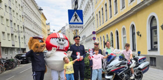 Eine Gruppe an Vertreter*innen der Polizei (Landespolizeivizepräsident General Dieter Csefan, interimistische Leiterin der Landesverkehrsabteilung Wien, Oberst Andrea Anders), Klaus Robatsch vom KFV, 3 Schulkindern mit Plüsch-HELMIS (1 davon auf einem Polizei-Motorrad) sowie den Maskottchen HELMI und dem Polizeibären vor einem Schild "Achtung Schulweg".