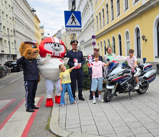 Eine Gruppe an Vertreter*innen der Polizei (Landespolizeivizepräsident General Dieter Csefan, interimistische Leiterin der Landesverkehrsabteilung Wien, Oberst Andrea Anders), Klaus Robatsch vom KFV, 3 Schulkindern mit Plüsch-HELMIS (1 davon auf einem Polizei-Motorrad) sowie den Maskottchen HELMI und dem Polizeibären vor einem Schild "Achtung Schulweg".