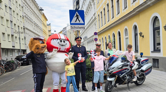 Eine Gruppe an Vertreter*innen der Polizei (Landespolizeivizepräsident General Dieter Csefan, interimistische Leiterin der Landesverkehrsabteilung Wien, Oberst Andrea Anders), Klaus Robatsch vom KFV, 3 Schulkindern mit Plüsch-HELMIS (1 davon auf einem Polizei-Motorrad) sowie den Maskottchen HELMI und dem Polizeibären vor einem Schild "Achtung Schulweg".