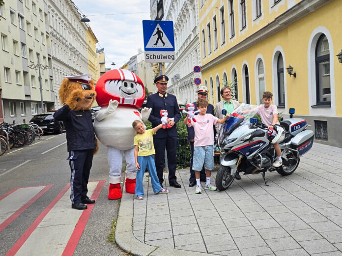 Polizei Aktion Schulanfang_Foto KFV Rosenkranz_02 Eine Gruppe an Vertreter*innen der Polizei (Landespolizeivizepräsident General Dieter Csefan, interimistische Leiterin der Landesverkehrsabteilung Wien, Oberst Andrea Anders), Klaus Robatsch vom KFV, 3 Schulkindern mit Plüsch-HELMIS (1 davon auf einem Polizei-Motorrad) sowie den Maskottchen HELMI und dem Polizeibären vor einem Schild "Achtung Schulweg".